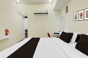 A hotel room with a bed, a desk, a chair, a red chair, a white vase with flowers, an air conditioning unit, and framed artwork on the wall. - Collection Kakadeo Kanpur Formerly Avon (Kanpur)