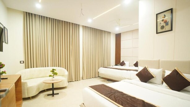 A modern hotel room with a bed, a sofa, a small round table, and a large window with curtains. - Hotel Shri Radha Krishnam (Barsana)