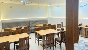 A restaurant with multiple tables and chairs, a cushioned bench seating area, pendant lighting, and a tall column structure. - Hotel Shri Radha Krishnam (Barsana)