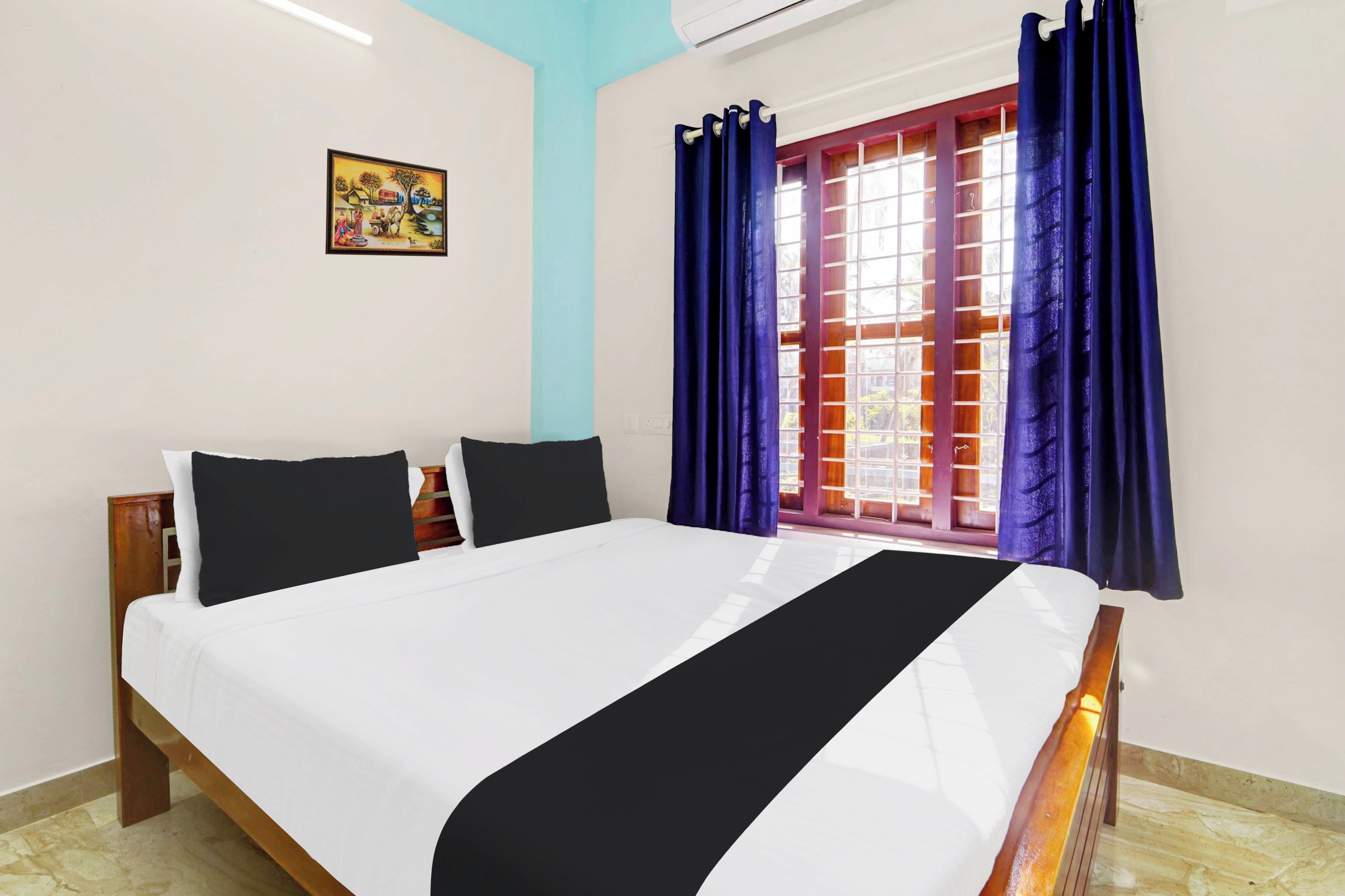 Une chambre à coucher avec un lit, deux oreillers, une table de chevet, une fenêtre avec des rideaux et un tableau accroché au mur.
