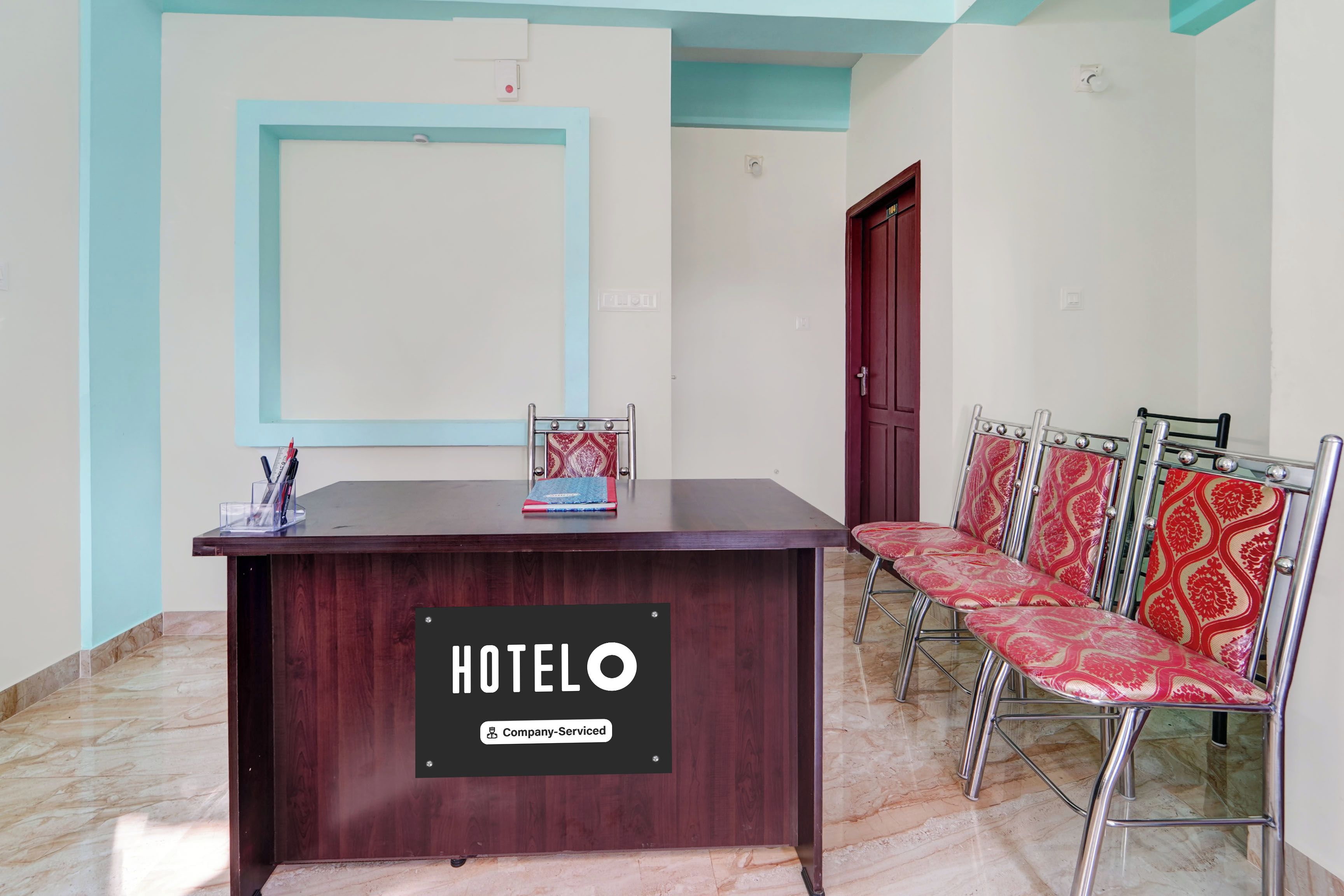 Un comptoir d’accueil d’hôtel avec une enseigne indiquant « HÔTEL O » et une étiquette « Service d’entreprise ». À droite, on aperçoit des chaises à armature métallique recouvertes d’un tissu à motifs.
