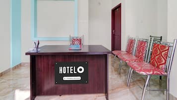 A hotel reception desk with a sign reading 'HOTEL O' and a 'Company-Serviced' label. There are metal-framed chairs with patterned upholstery to the right.