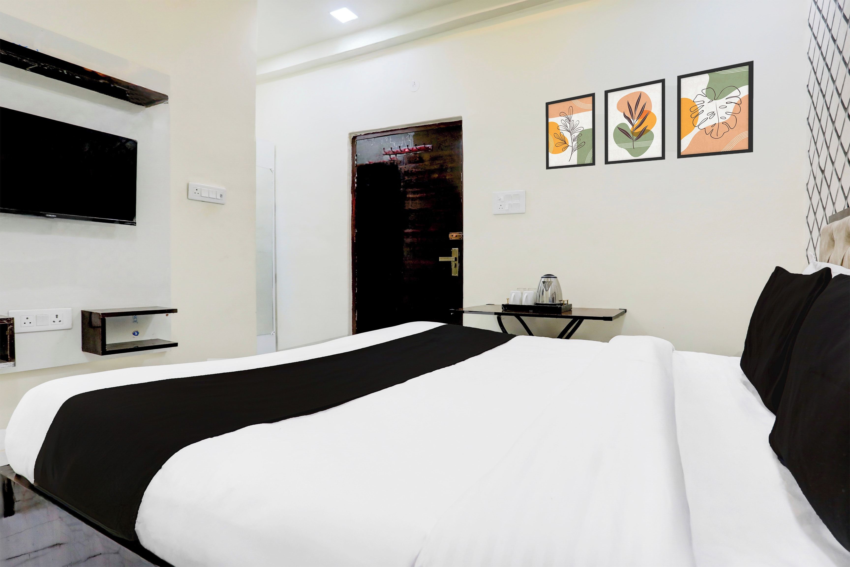 A hotel room with a large bed, a flat-screen TV mounted on the wall, a small table with a kettle, and three framed artworks on the wall.