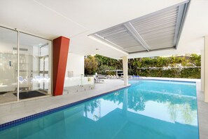 Ein moderner, überdachter Innenpool mit klarem, blauem Wasser, einer Glaswand und Liegestühlen.