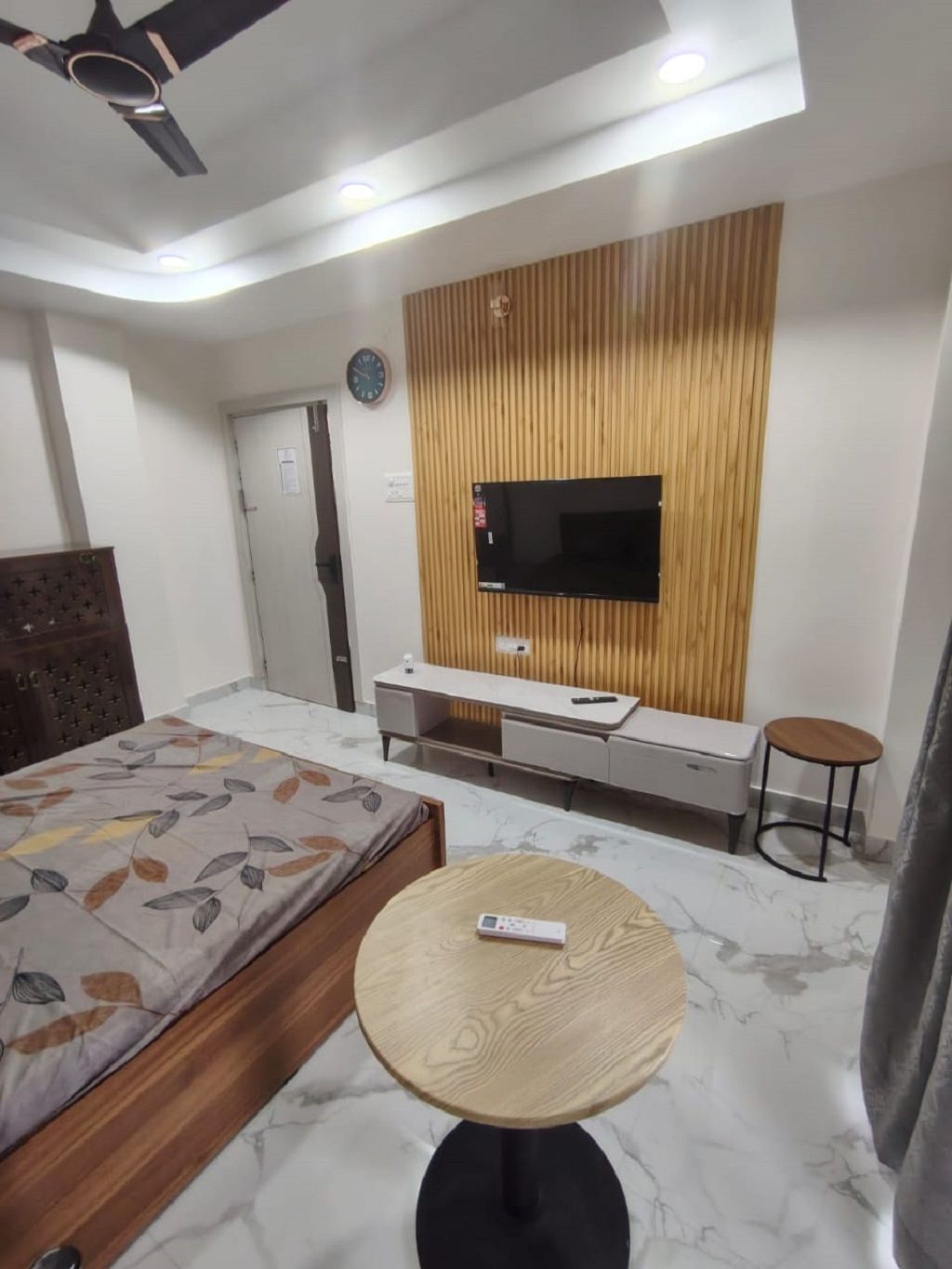 Quarto de hotel moderno com uma cama, uma mesa redonda de madeira, uma TV montada em painel de parede de madeira, uma escrivaninha pequena e uma cadeira.