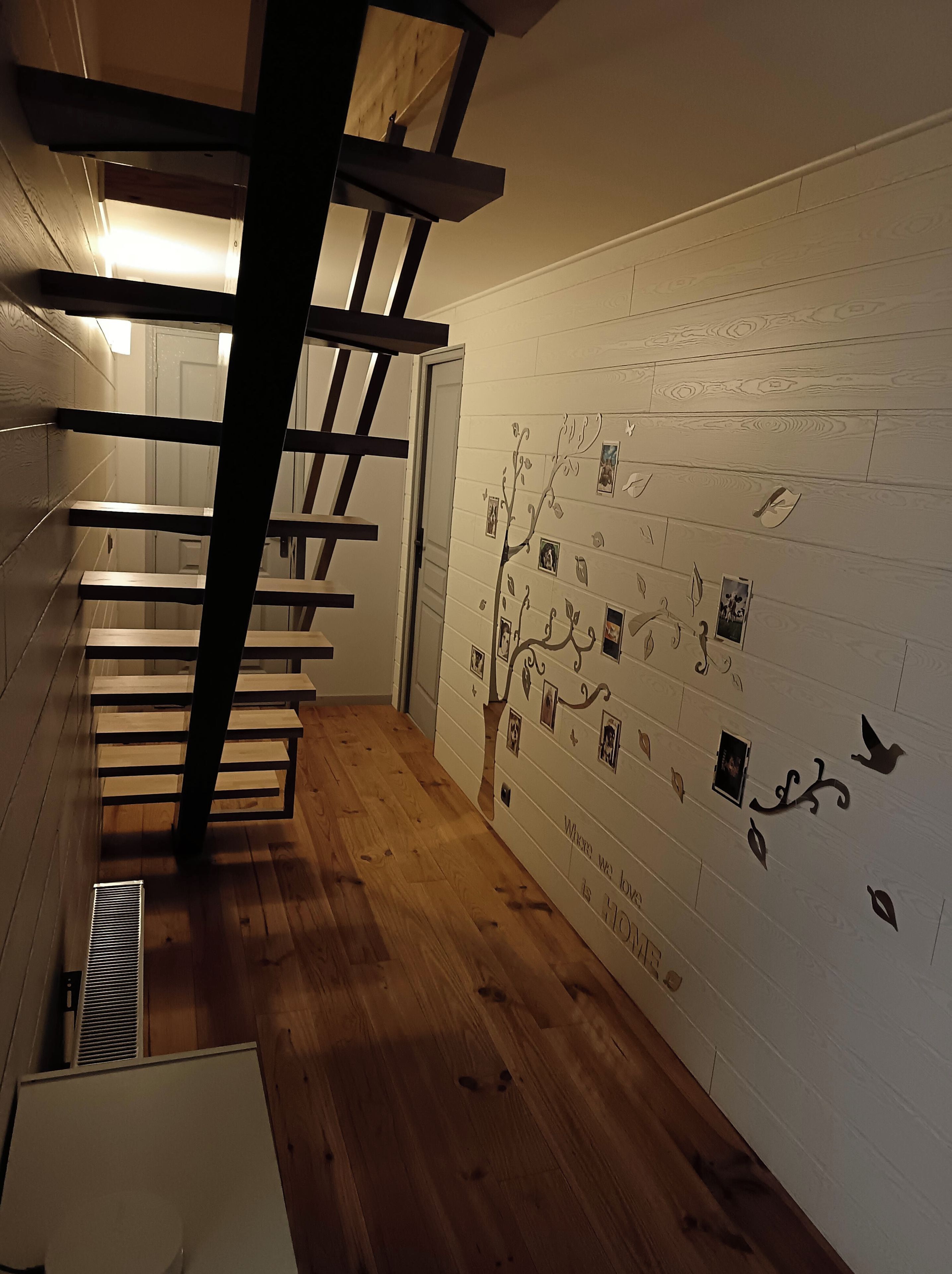 Een trap met houten treden en een metalen leuning, die leidt naar een kamer met witte wanden versierd met ingelijste foto's en een muurschildering van een boom.