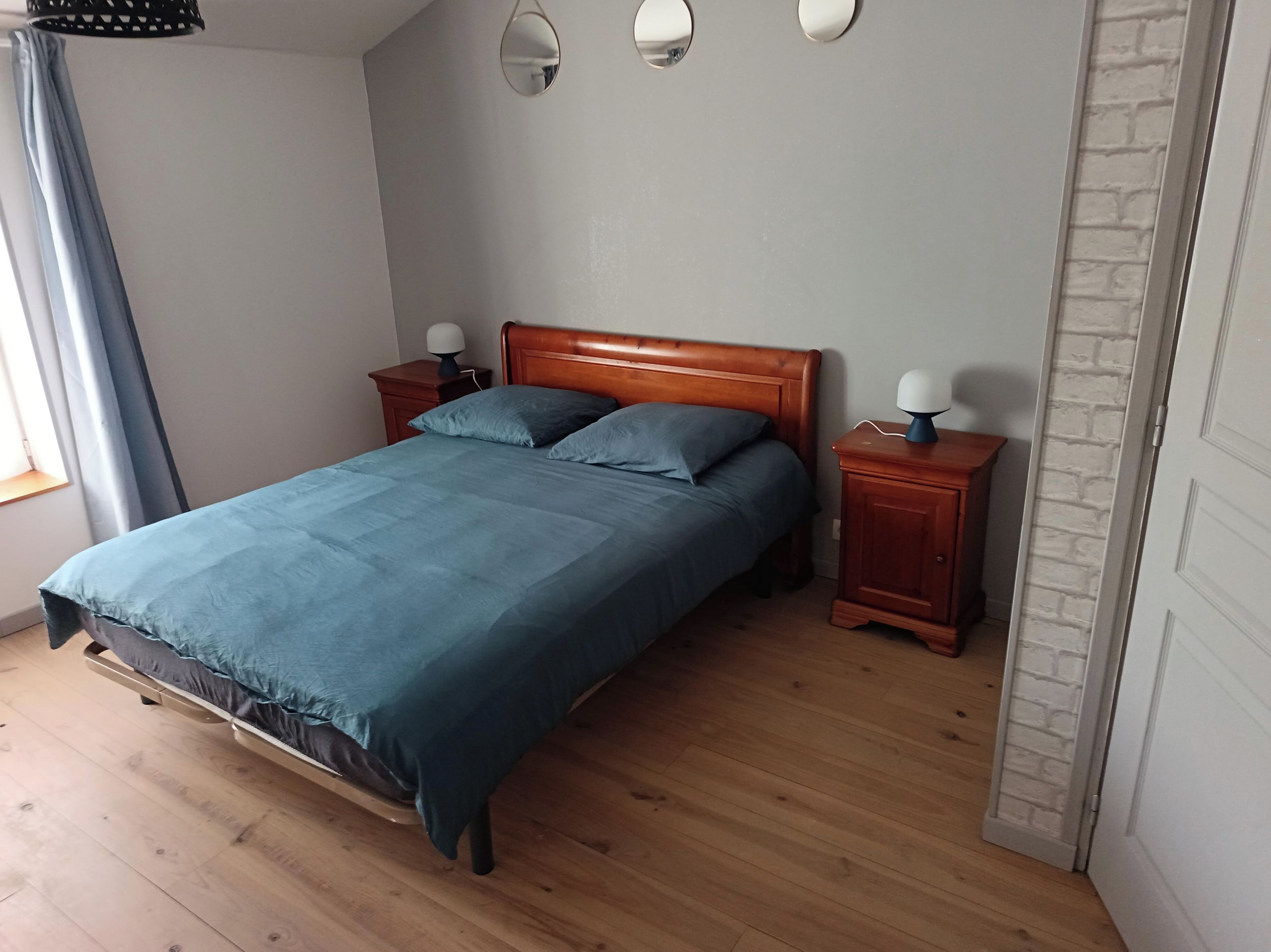 A bedroom with a wooden bed, two bedside tables with lamps, a window with curtains, and a brick-patterned wall.