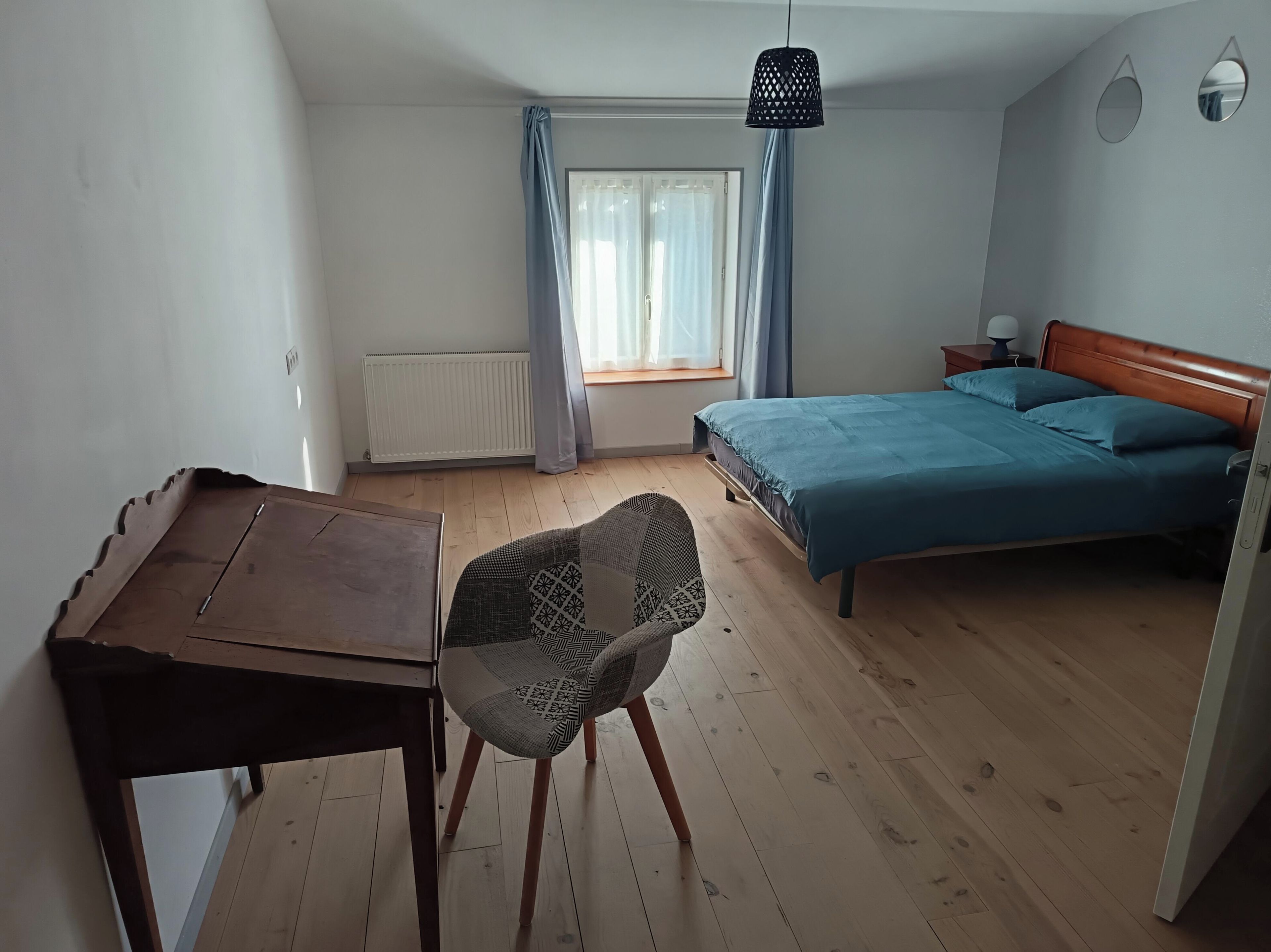 Une chambre à coucher comprenant un lit, une chaise, une table, une fenêtre avec des rideaux, un radiateur et un luminaire suspendu.