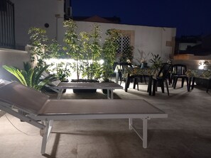 En udendørs patio med liggestol, spisebord, stole og krukker med planter.