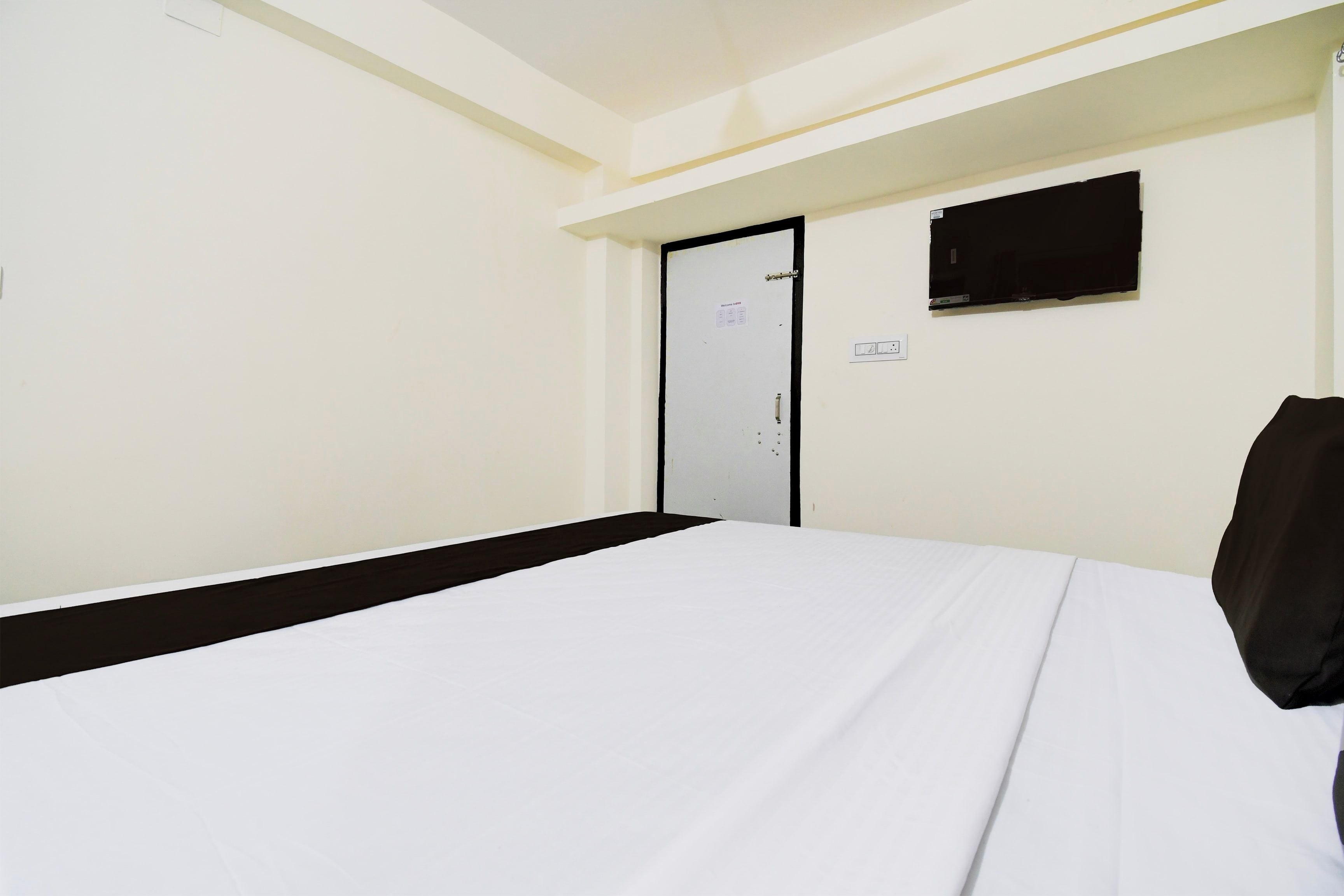 Habitación de hotel con cama, puerta, televisor montado en la pared y toma de corriente.