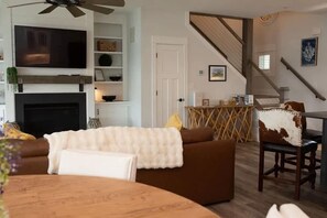 A living room with a fireplace, a television mounted above it, a brown sofa, a wooden coffee table, and a dining area with a table and chairs. - Sea La Vie House wPool | The Inn on Turville Creek (Berlin)