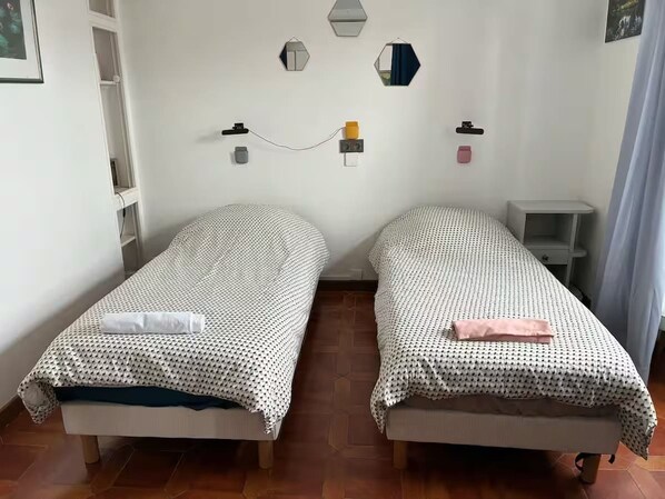 Habitació amb dos llits individuals, una taula petita i una estant mural amb diversos objectes.