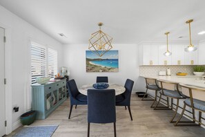 A modern kitchen with a dining area, featuring a round table with chairs, a pendant light, and a painting of a coastal scene. - Block from the Beach – Pristine Avalon Condo Just One Block from the Beach! (Avalon)