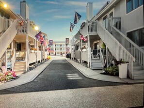 A street lined with two-story buildings with balconies, a clear sky, and flags. - Block from the Beach – Pristine Avalon Condo Just One Block from the Beach! (Avalon)