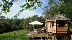 Une cabane en bois rustique avec une terrasse, un parasol blanc et un véhicule garé à proximité.