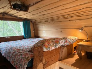 Quarto de cabana com cama, escrivaninha, luminária e janela com cortinas.