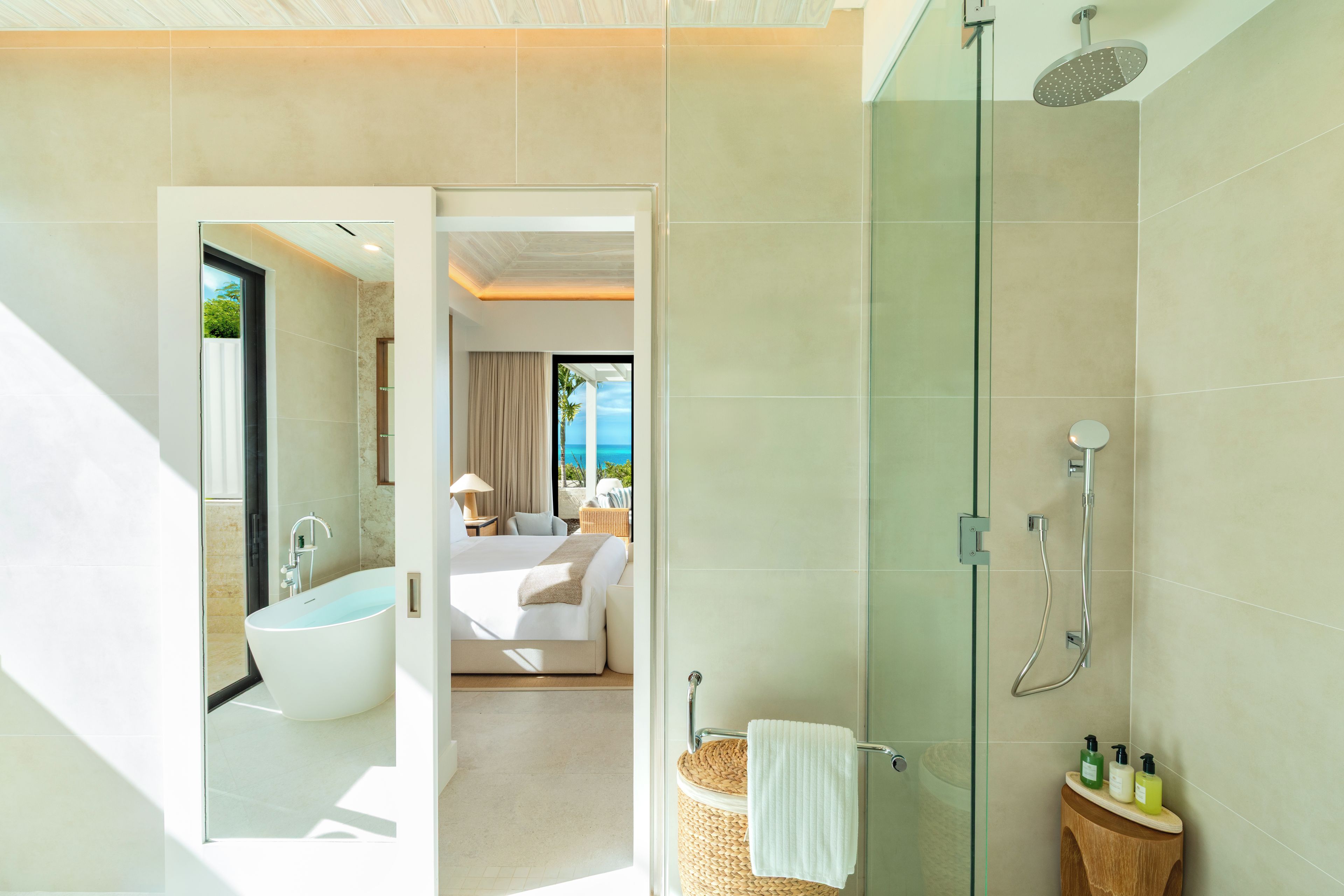 Un baño moderno con cabina de ducha de vidrio, bañera y una habitación visible con cama y cortinas.