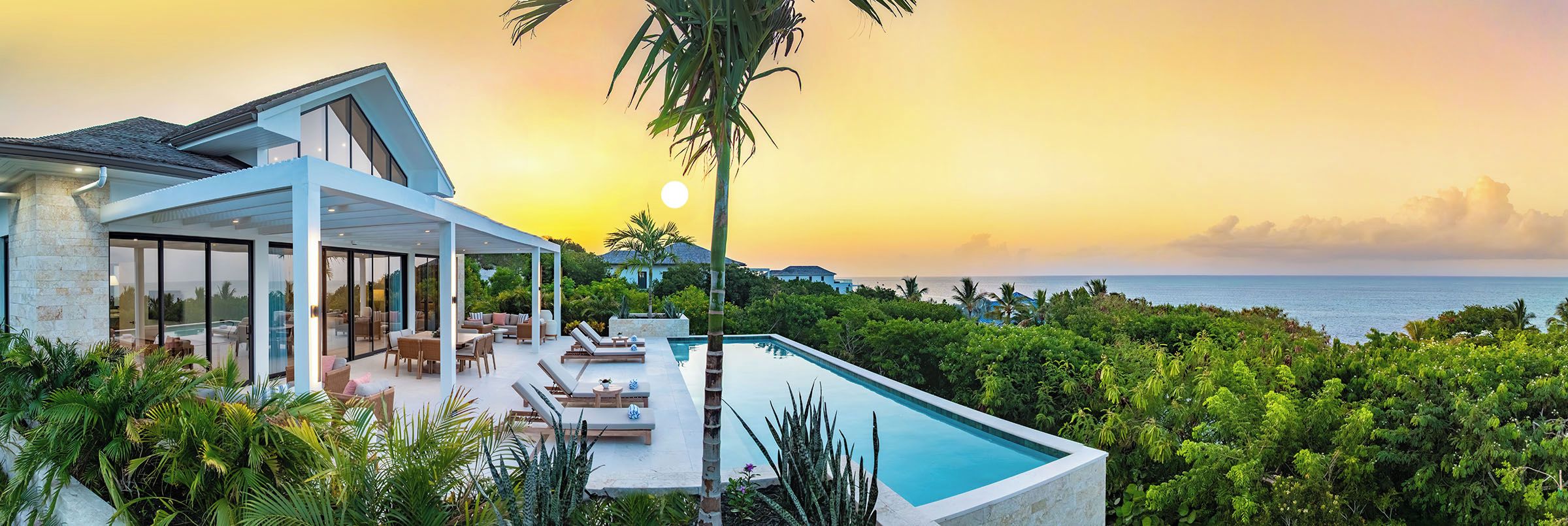 Una casa moderna con piscina, mobiliario exterior y vista al océano al atardecer.