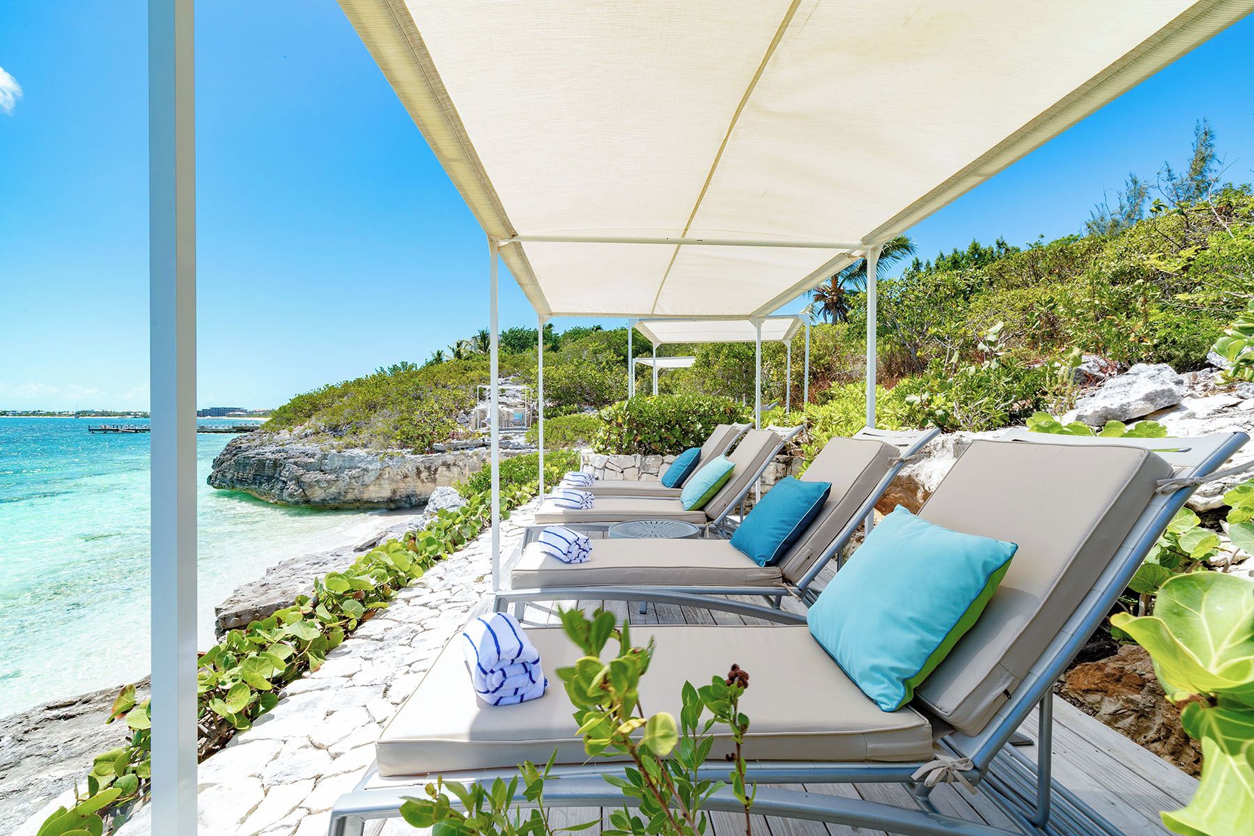 Un espacio de estar al aire libre techado con sillones reclinables y sombrillas, con vista a una playa de aguas cristalinas y terreno rocoso.