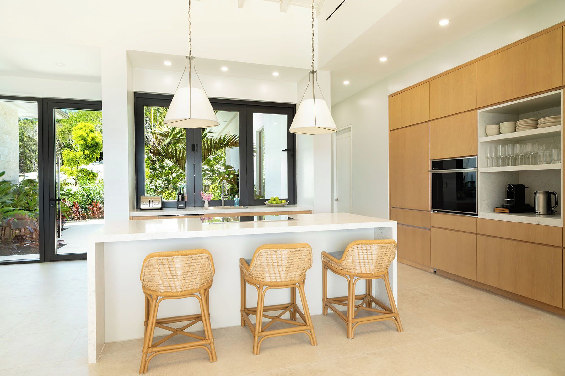 Una cocina moderna con una isla central, tres sillas de mimbre, iluminación colgante y vistas a zonas verdes al exterior.