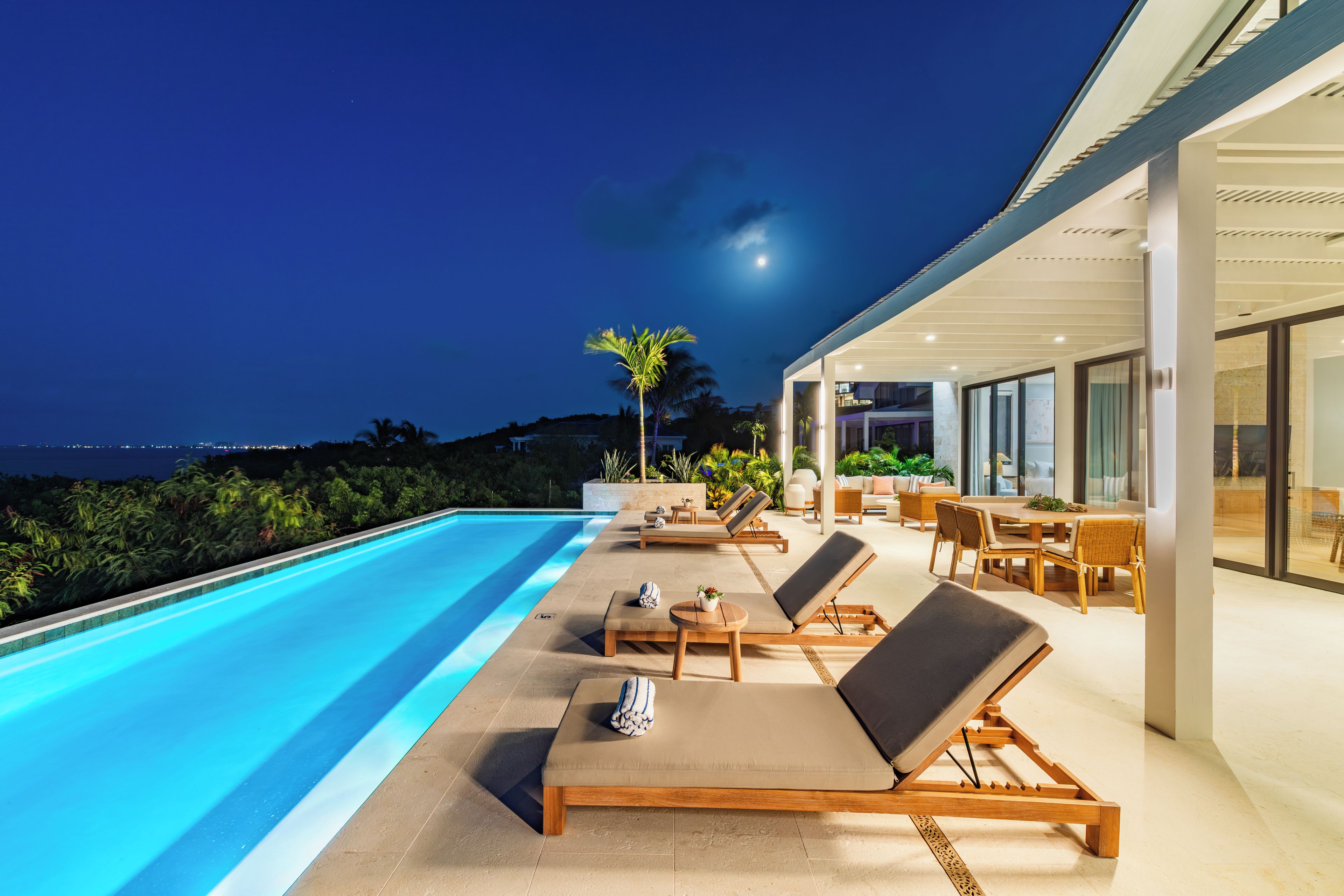 Área de piscina com espreguiçadeiras e mesa de jantar, com vista para um corpo d'água à noite.
