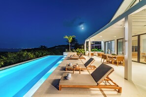 A poolside area with lounge chairs and a dining table, overlooking a body of water at night.