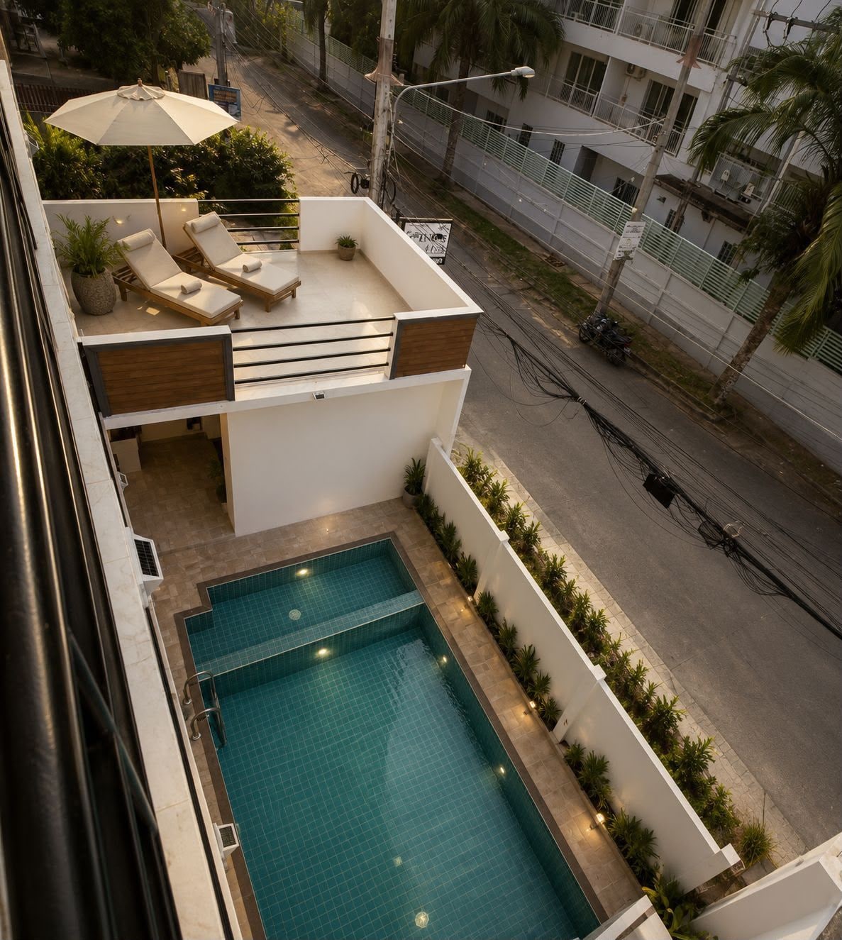 A rooftop pool area with lounge chairs and an umbrella, surrounded by a white wall and plants.