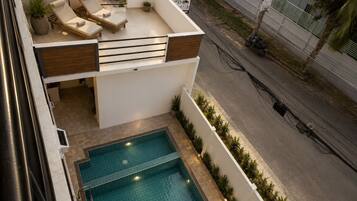 Uma área de piscina no terraço com espreguiçadeiras e um guarda-sol, cercada por um muro branco e plantas.