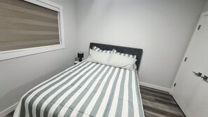 A small, single-bed room with a striped bedspread, a bedside table with a lamp, and a window with blinds. - Cozy Laneway Suite in Harbour Landing (Regina)