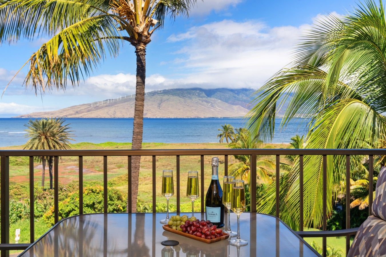 A balcony with a table set for two, overlooking a coastal landscape with palm trees and a view of the sea and mountains.