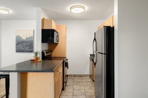 A compact kitchen with wooden cabinets, a black microwave, a stainless steel refrigerator, and a granite countertop. - Sweeping Ocean and Mountain Views from Every Room! (Kihei)