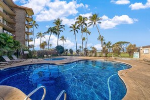 A swimming pool with a curved edge, surrounded by palm trees and a building with balconies. - Sweeping Ocean and Mountain Views from Every Room! (Kihei)