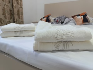 Cama bem arrumada com roupa de cama branca e toalha dobrada com desenho de palmeira.