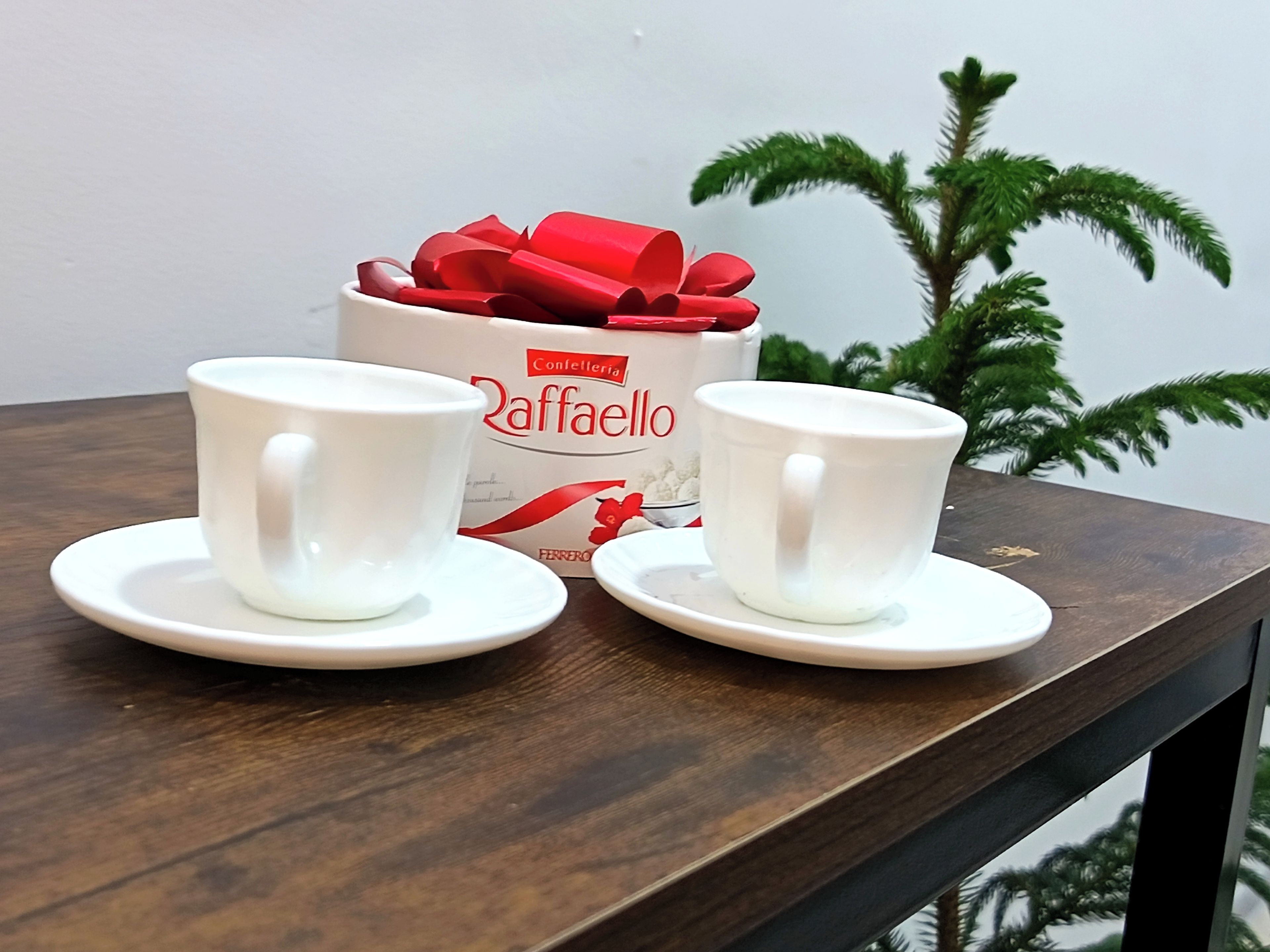 Dos tazas blancas sobre platillos en una mesa de madera, con un recipiente blanco al fondo.