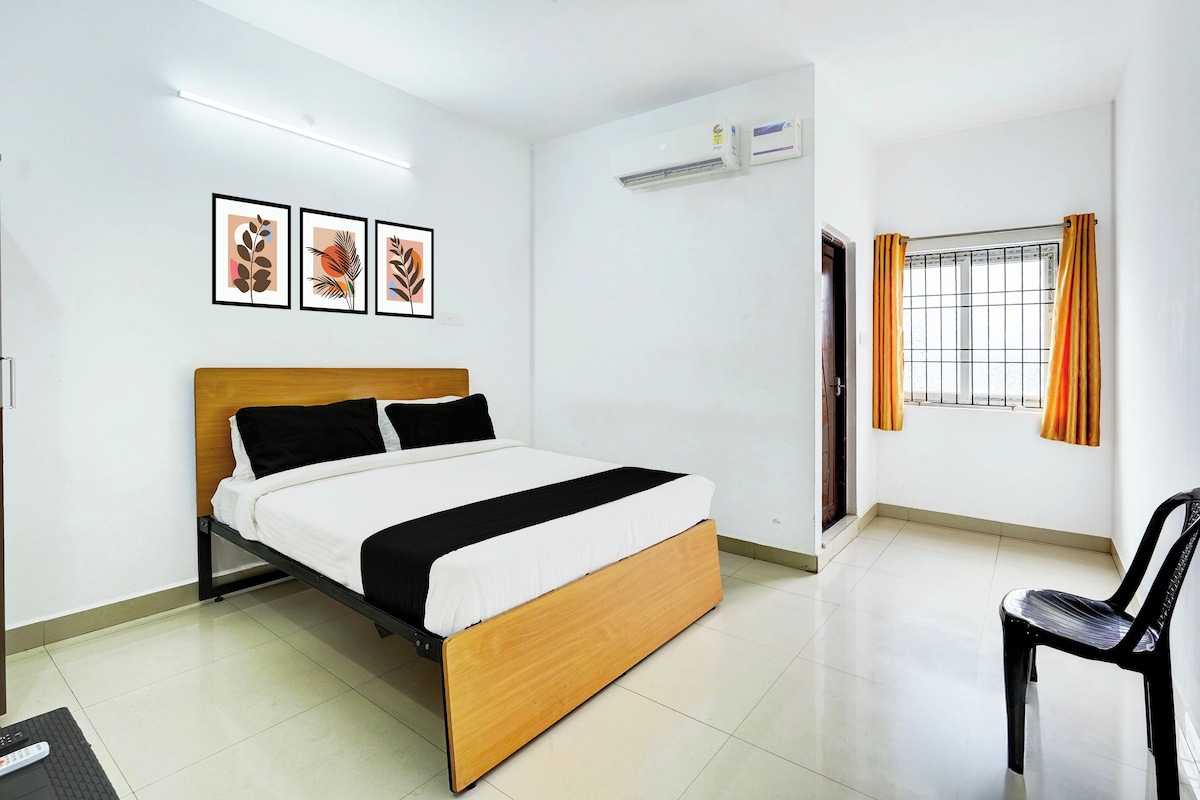 A hotel room with a bed, a chair, an air conditioning unit, a window with curtains, and framed artwork on the wall.