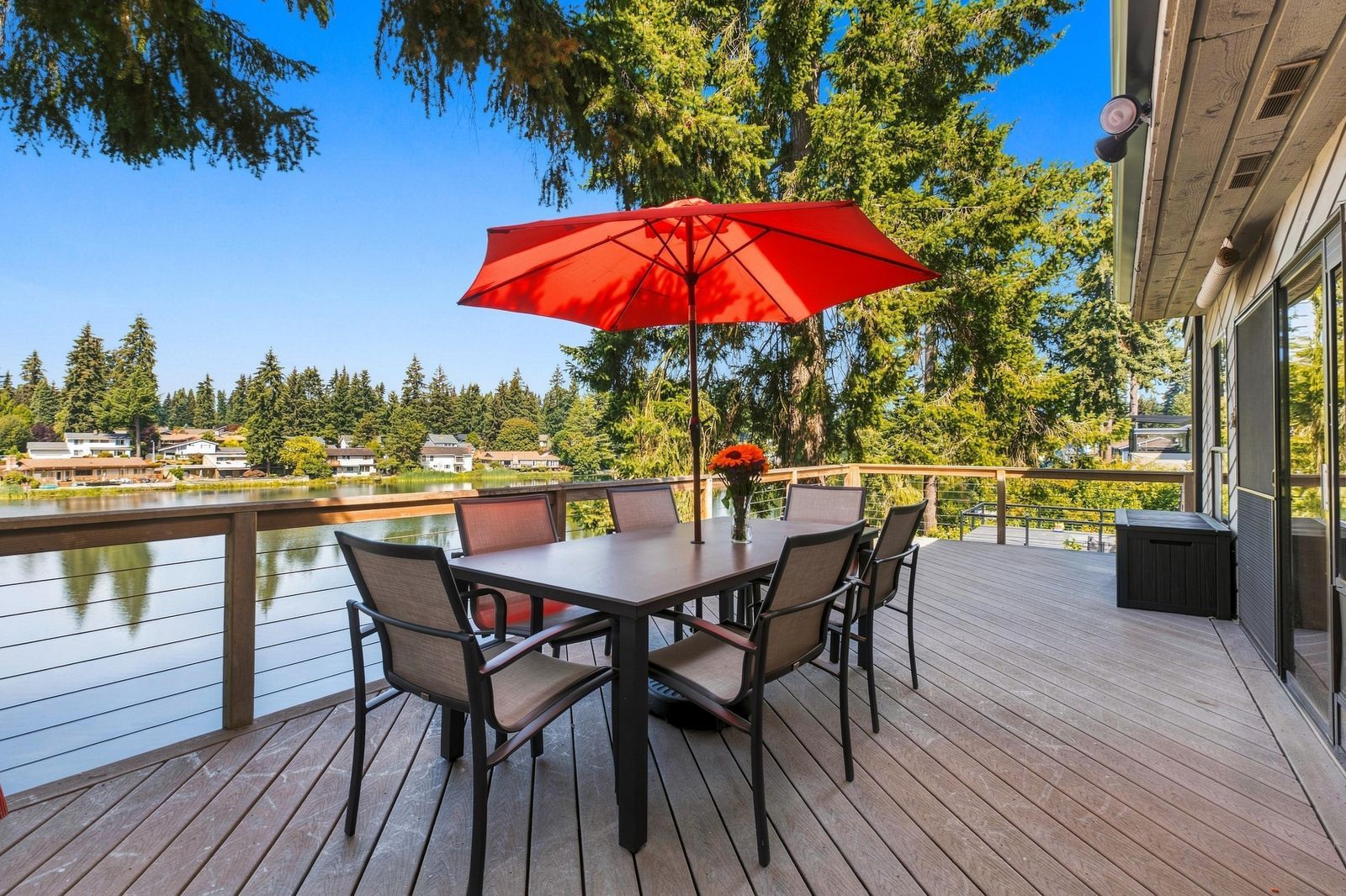Um terraço com mesa e cadeiras, um guarda-sol vermelho e vista para um lago e árvores.