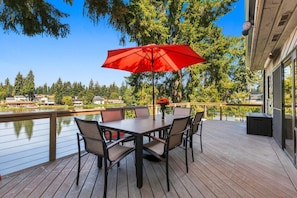 A deck with a table and chairs, a red umbrella, and a view of a lake and trees. - Lake House Retreat w/Sauna, golf course, 30 min 2 Seattle, game room, water toys (Federal Way)