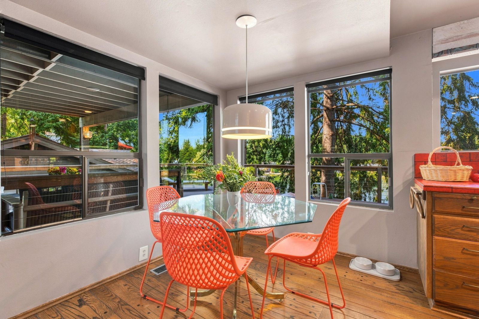 Un coin repas avec une table en verre, des chaises orange et de grandes fenêtres donnant sur un paysage arboré et une terrasse.