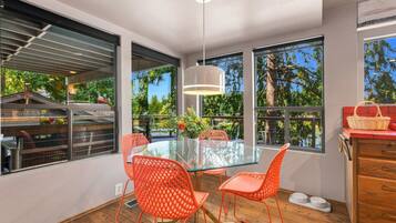 Un coin repas avec une table en verre, des chaises orange et de grandes fenêtres donnant sur un paysage arboré et une terrasse.