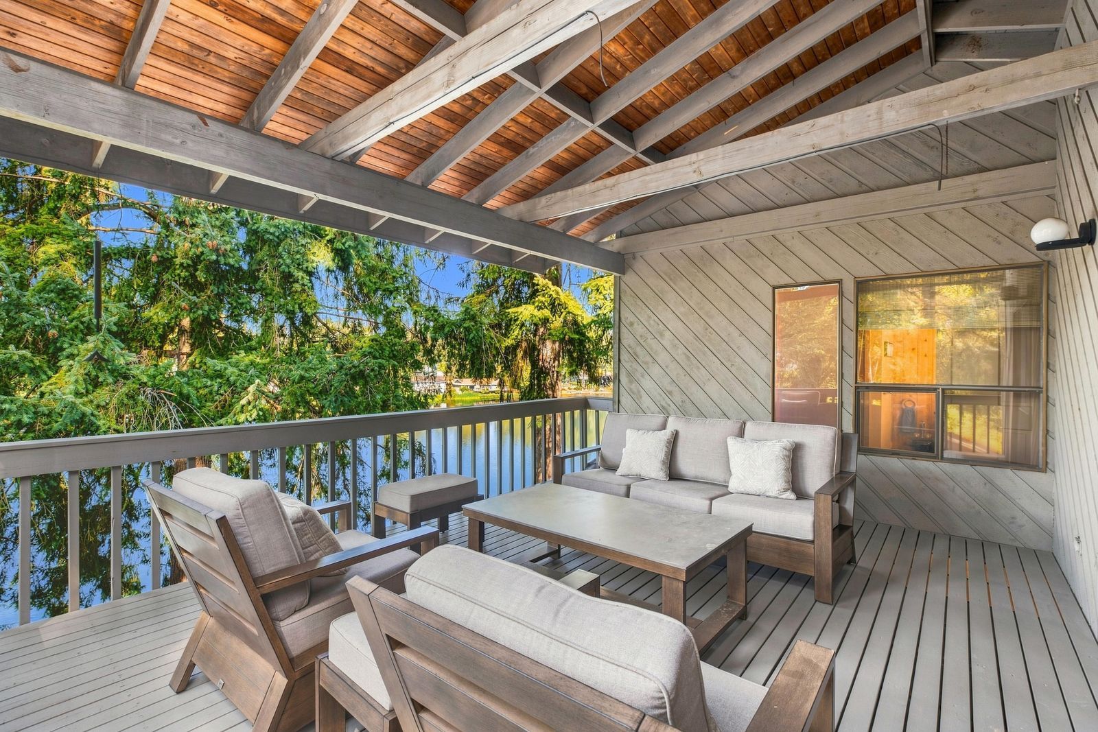 A covered outdoor deck with wooden furniture, a view of the water, and a large window.
