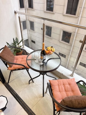 Un balcon avec une table en verre, deux chaises, une plante en pot, une bouteille de champagne et un panier de fruits.