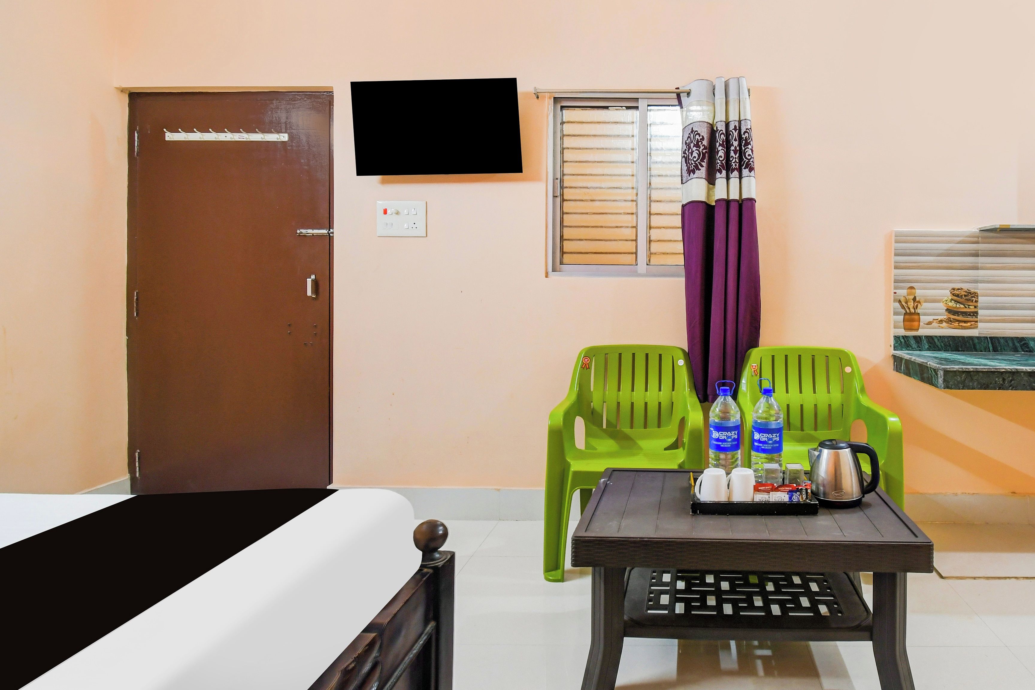 Habitación de hotel con cama, una mesa con hervidor, dos sillas, ventana con persianas y televisión en la pared.