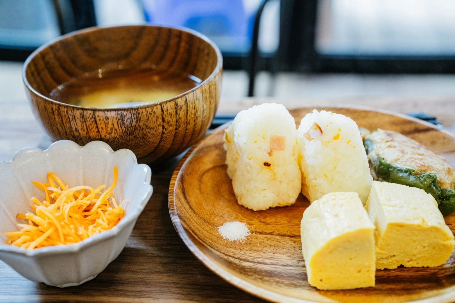 A wooden table with a bowl of soup, a plate with rice balls, shredded cheese, and a green vegetable.