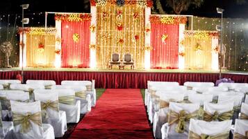 Un escenario con cortinas rojas y adornos florales, dos sillas y una alfombra roja que conduce a sillas blancas con fajines dorados.