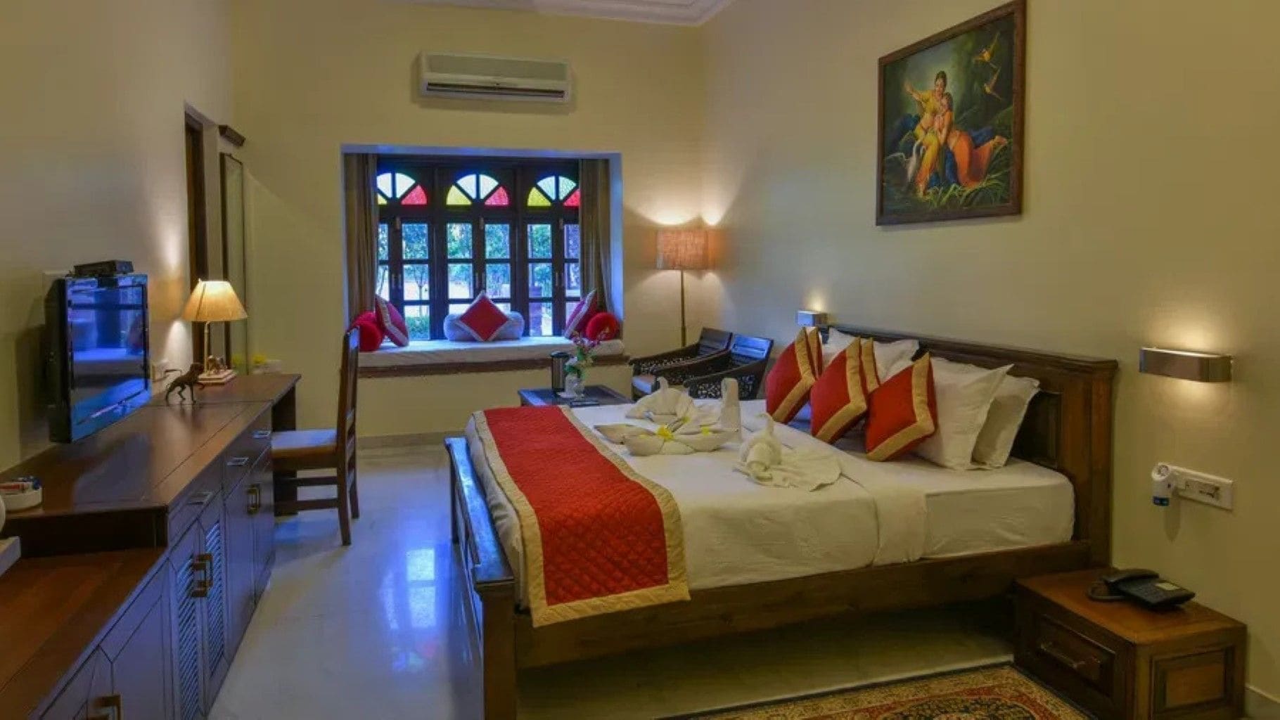 A hotel room with a bed, a desk, a television, a window with stained glass, and a painting on the wall.