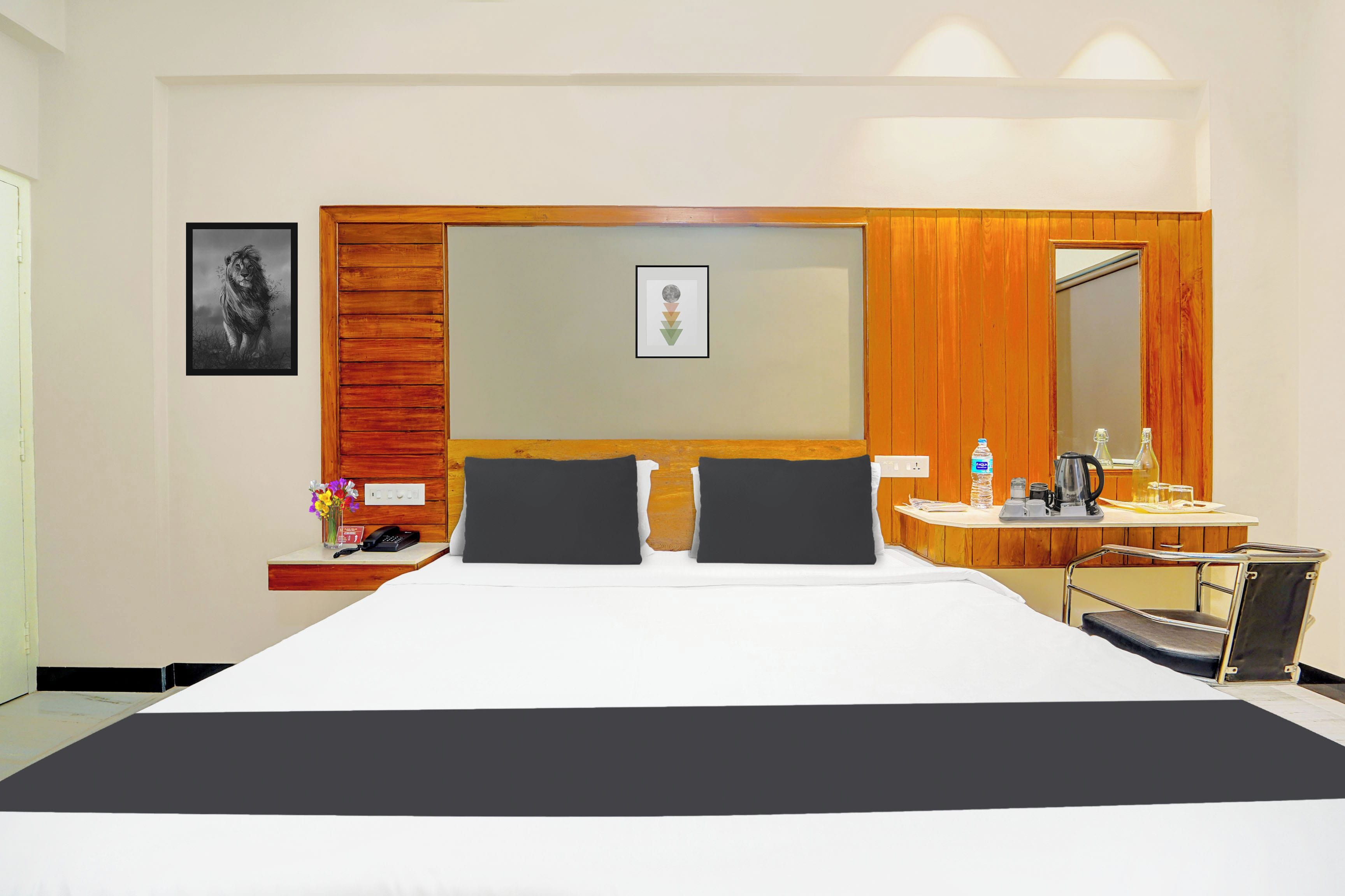Een hotelkamer met een groot bed, een nachtkastje met een telefoon en een bloevas, een spiegel en een bureau met een waterkoker en een fles water.