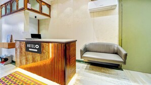 A hotel reception area with a wooden reception desk, a grey sofa, and a wall-mounted air conditioner.