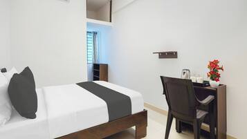 Une chambre d’hôtel avec un lit, un bureau et une chaise. Il y a une fenêtre avec des rideaux, une plante et un vase rempli de fleurs.