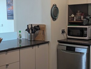 A kitchen with a microwave, dishwasher, and a wooden cutting board with utensils. - Fantastic sunny arty apartment, fully equipped and furnished for short let (Southbank)