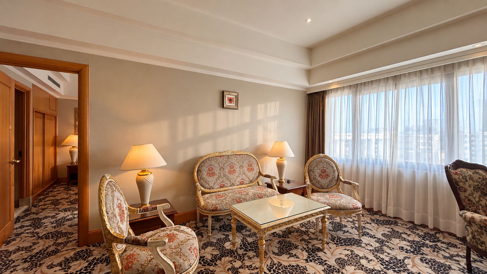 Habitació d'hotel amb zona de seients, incloent dues cadires i una taula, un gran finestral amb cortines i una porta que dona a una altra habitació.
